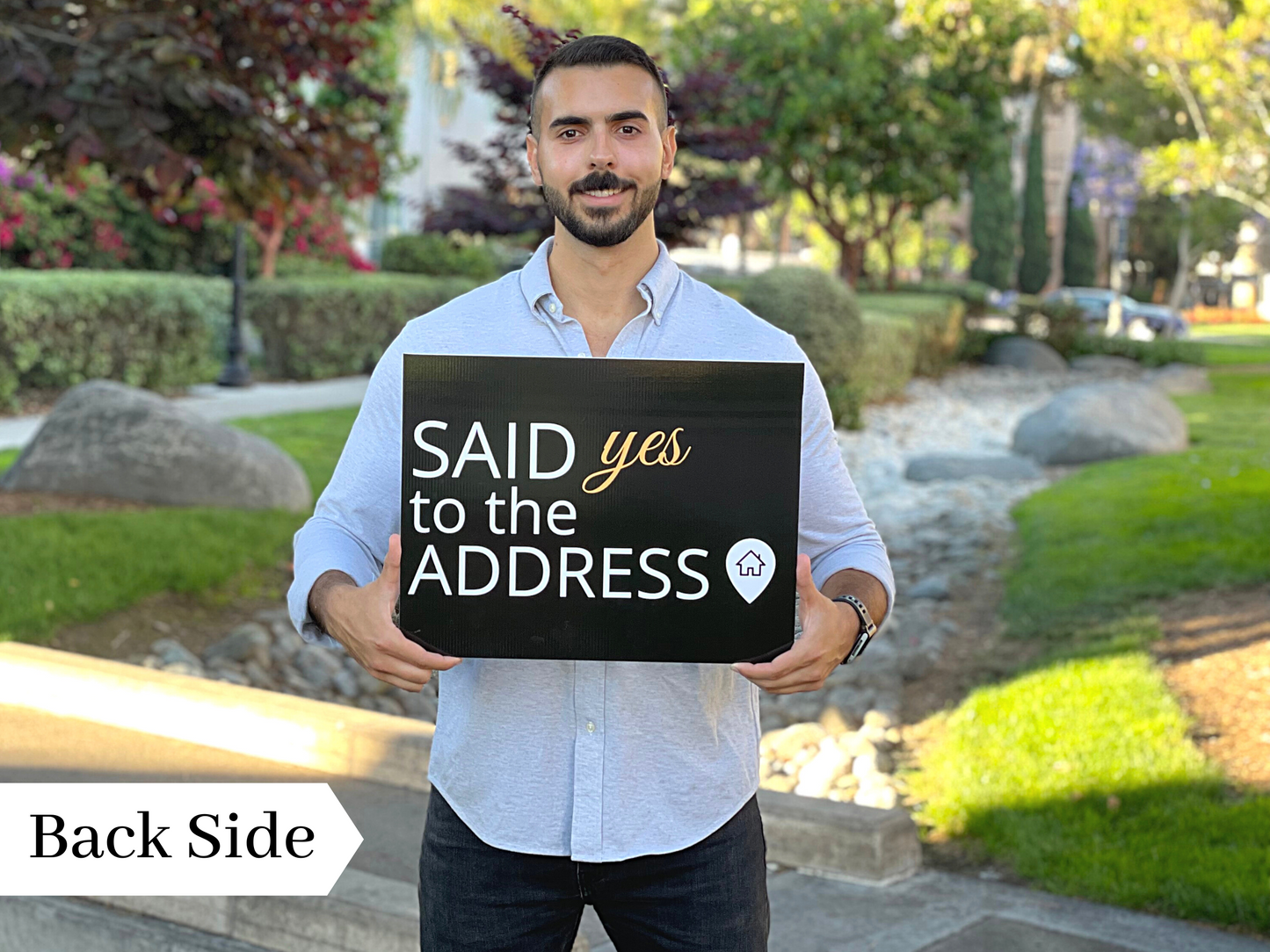 Realtor holding the back side of a rectangle Said Yes to the Address prop sign in a garden setting