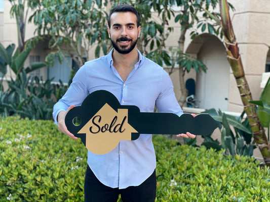 Realtor smiling and holding a double-sided key-shaped Sold prop sign outdoors near a building